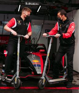 two racecar drivers standing on shell ride electric scooters in a garage